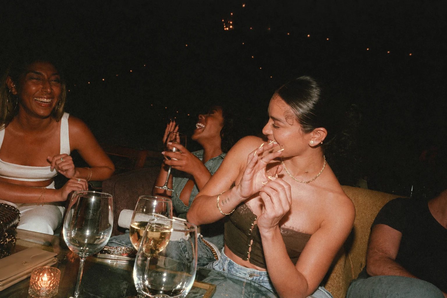 Friends laughing around a low table late at night, with wine glasses and a candle in front of them and string lights overhead.